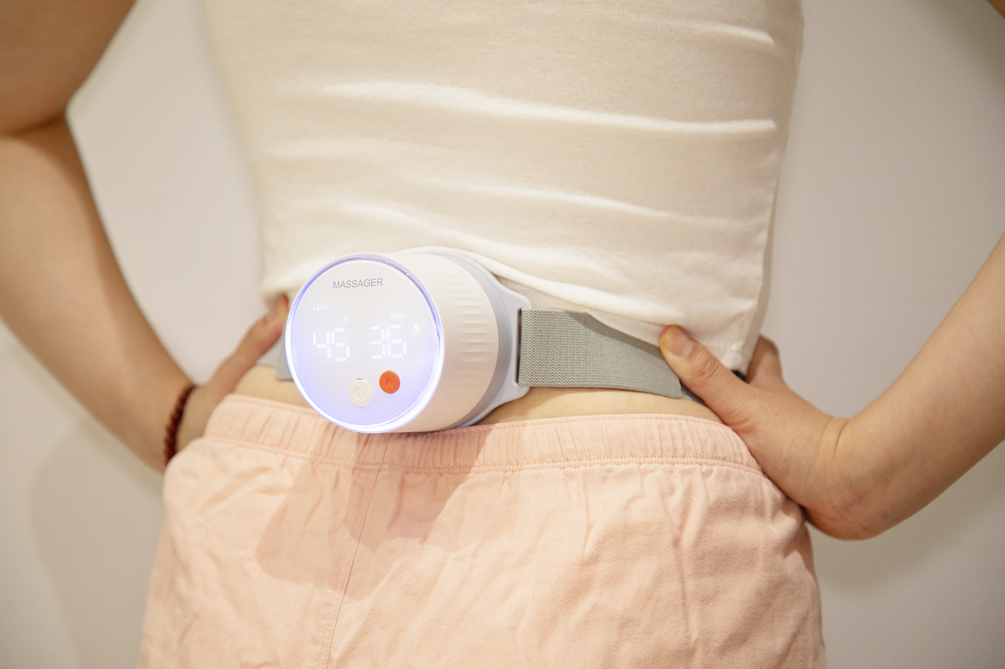 Woman wearing a white cotton top and pink loose pants using a smart moxibustion device on her lower back in a cozy home setting with warm lighting.